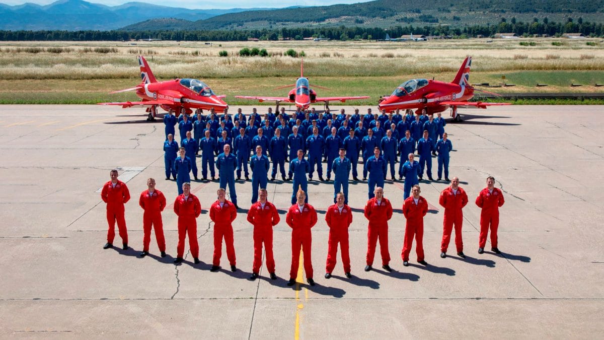 Royal Air Force (RAF) members wearing red and blue stand in formation on an air field