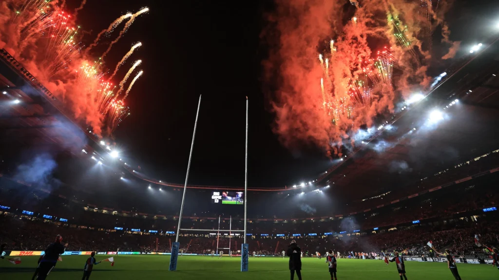 Rugby stadium at night with full crowd and fireworks exploding overhead