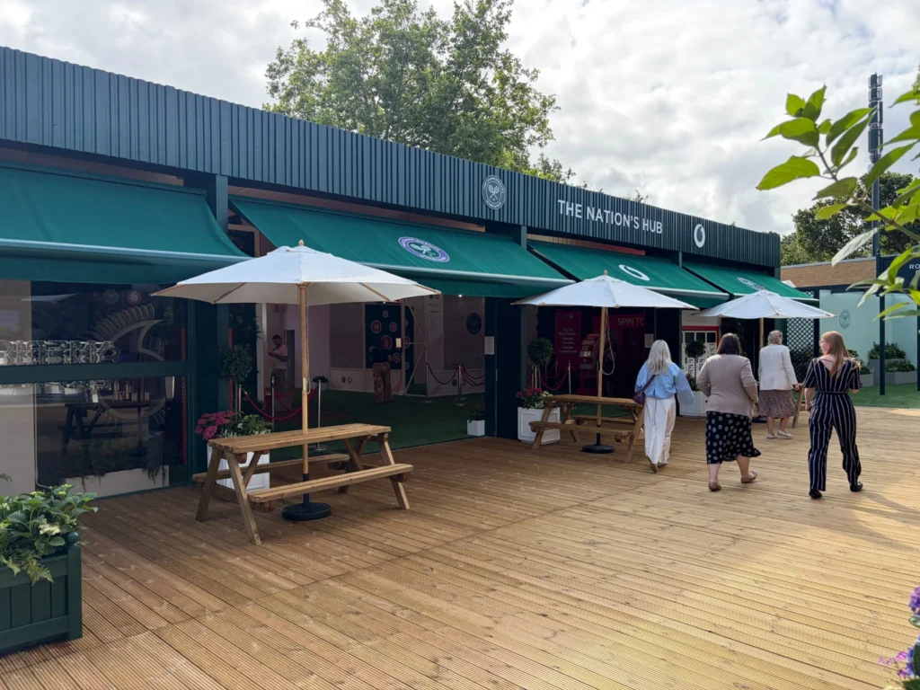 Exterior of The Nation's Hub, Vodafone's fan experience space at Wimbledon 2026, with green awnings and picnic benches